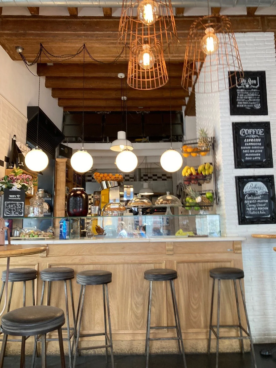 Interior of a coffee shop with hanging light fixtures, a counter with a glass display case, chairs, and a menu board on the wall listing coffee and bakery items.
