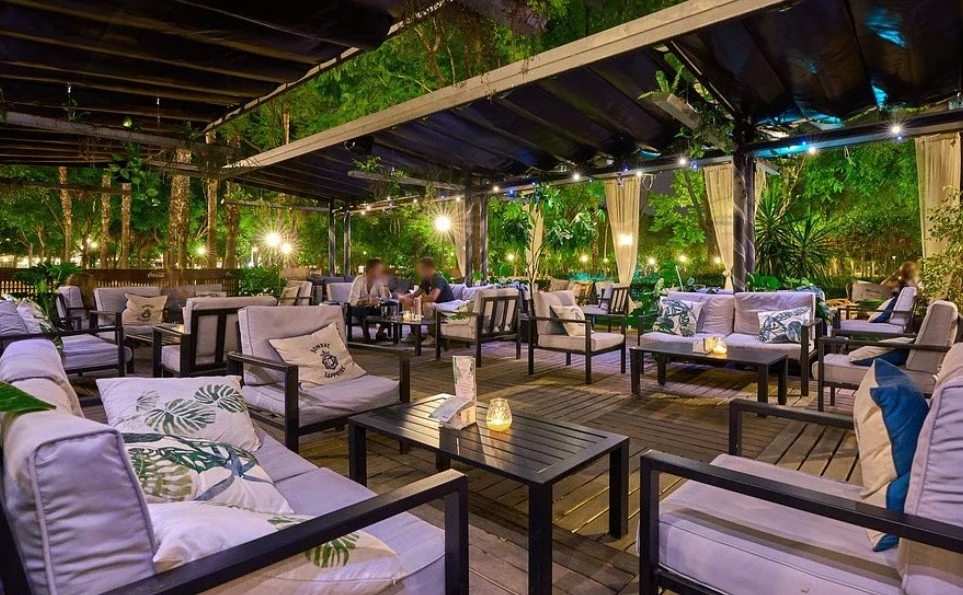 An outdoor restaurant or lounge area at night with comfortable seating, pillows, small tables with candles, surrounded by lush green trees and plants.