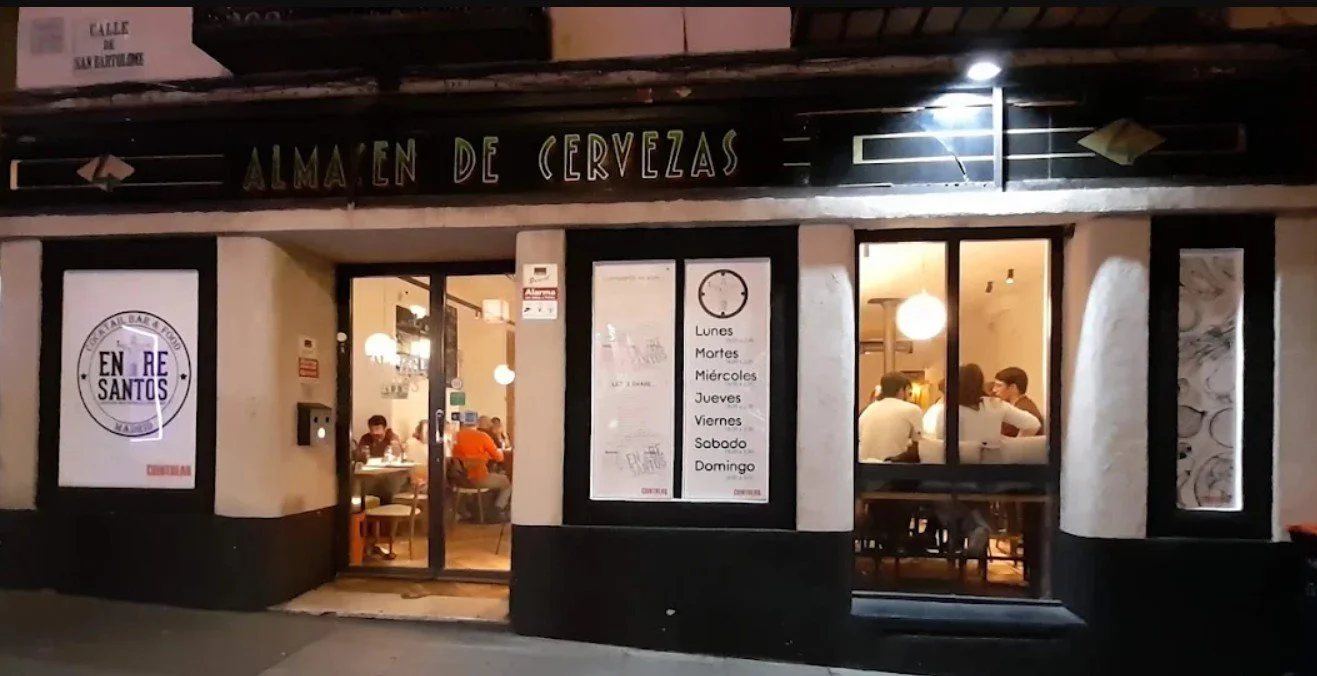 Exterior of a restaurant with glass windows showing customers dining inside, and signage indicating the restaurant is an ale house. The sign above reads 'Alma en de cervezas.' There are menus listing days of the week and a large poster on the left side with the restaurant's logo and name.