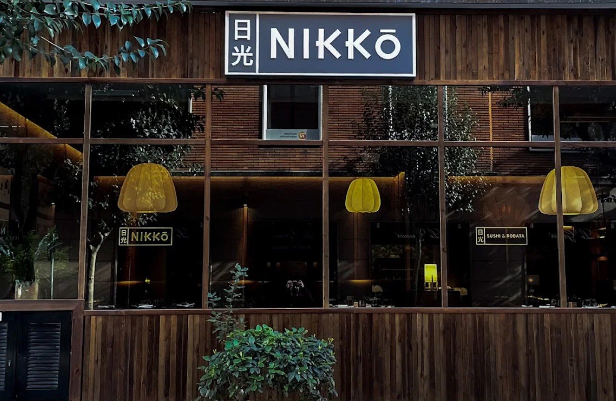 Exterior of a restaurant named NIKKO with large glass windows, wooden framing, and yellow pendant lights inside. There are plants outside and Japanese characters on signs.