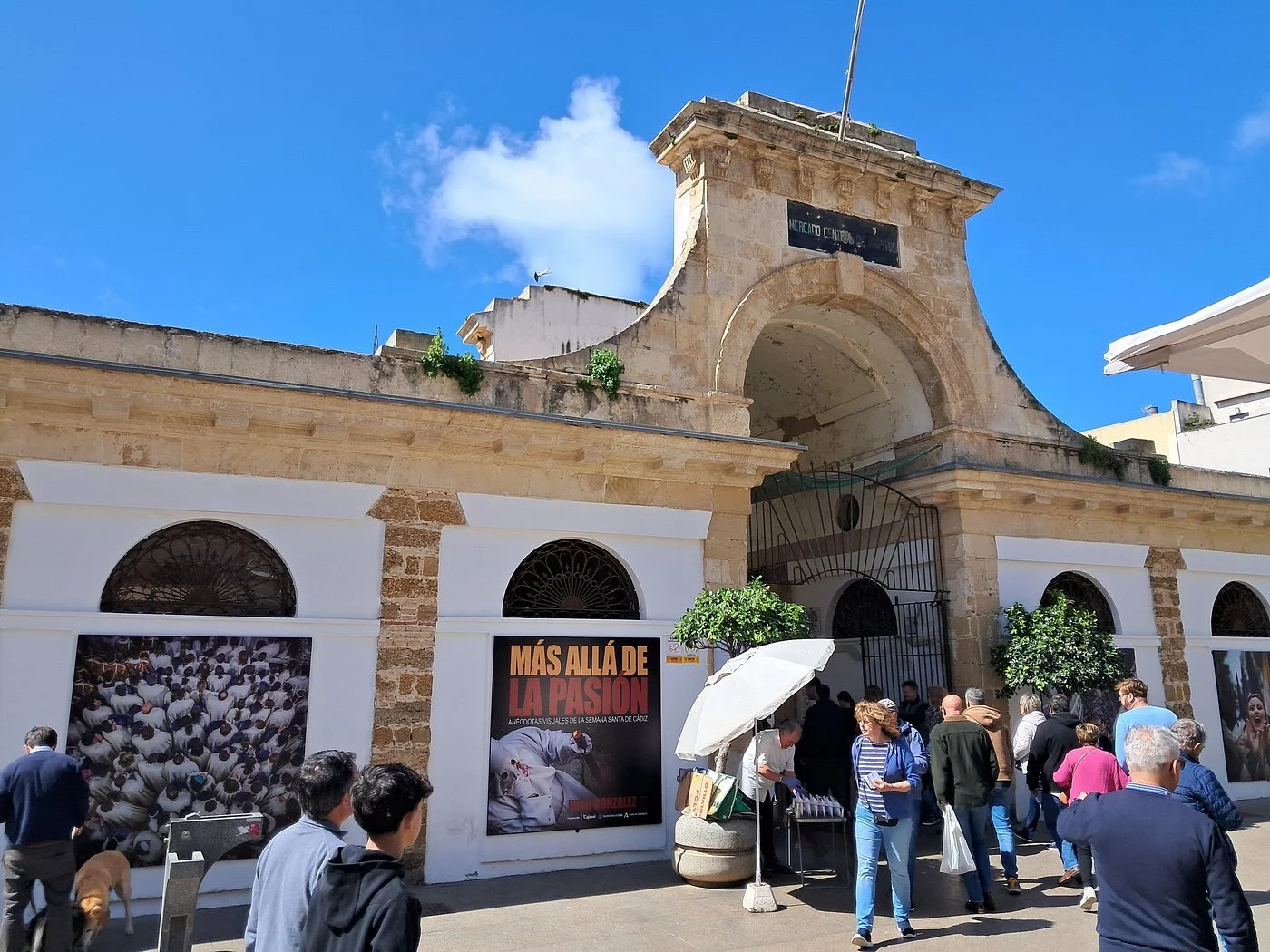 A historical stone archway entrance with a black sign and gate, people walking and standing outside in a plaza, bright blue sky with a few clouds, advertisements on the walls, and a parasol providing shade.