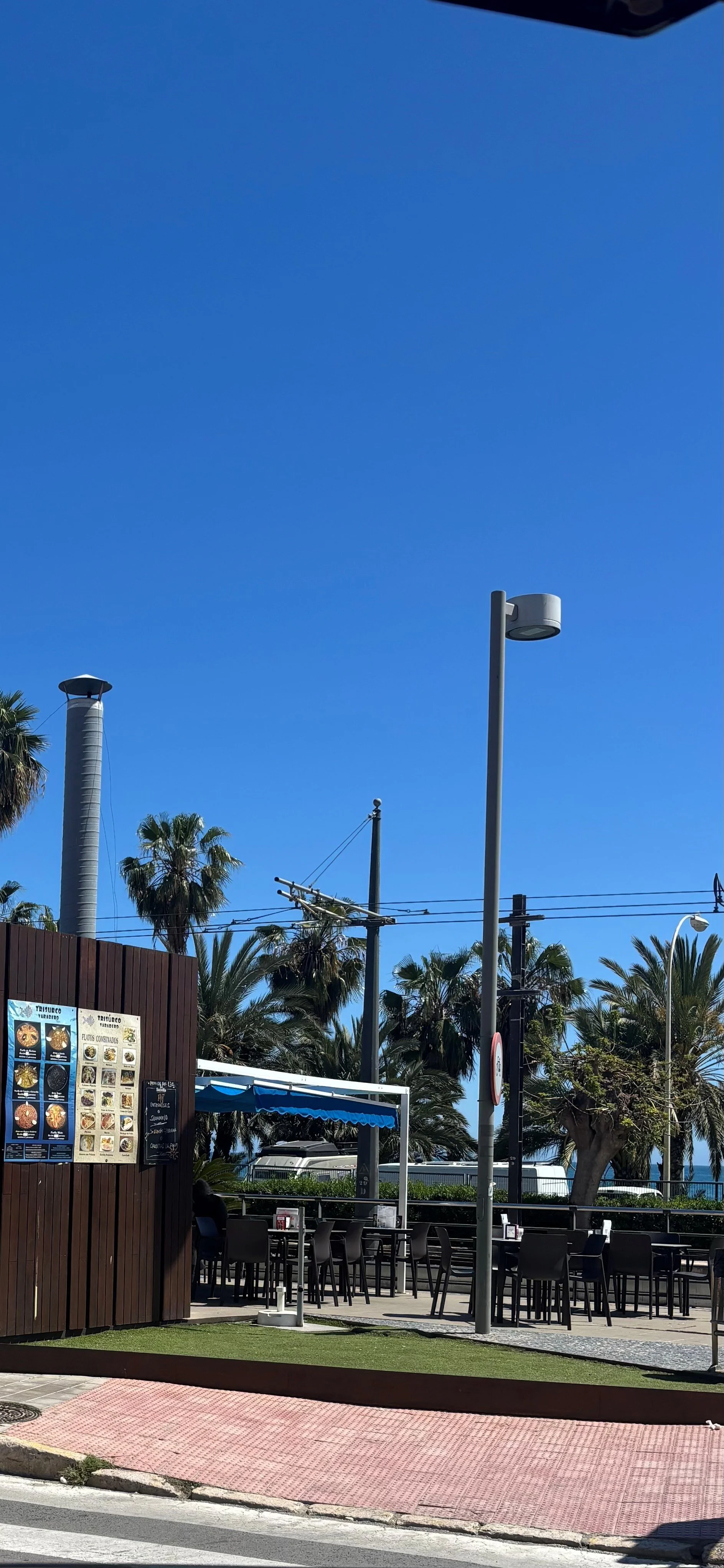 Outdoor cafe with stacked chairs and tables, menu board, palm trees, utility poles, and a clear blue sky.