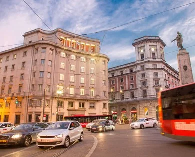 City street scene with historic buildings, cars, a red bus, and a statue on a pedestal during dusk.