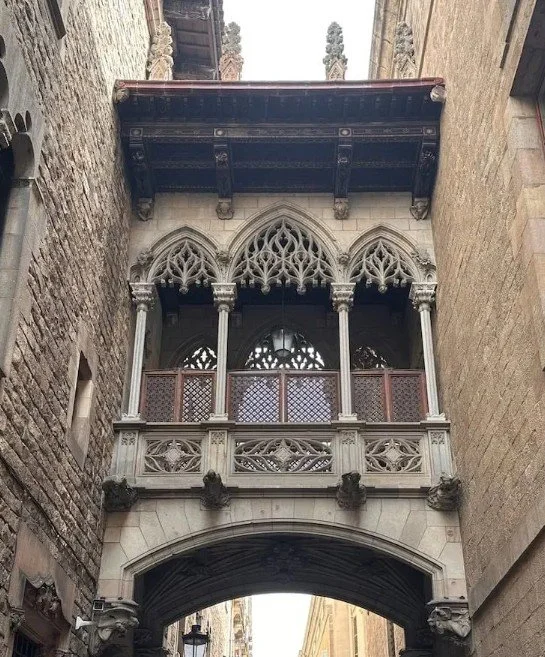 Gothic-style architectural balcony with pointed arches, decorative tracery, and stone columns, extending over a narrow street between stone buildings.