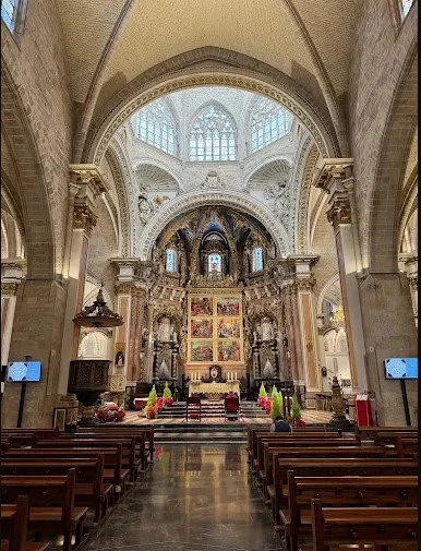 Interior view of a grand church or cathedral with high vaulted ceilings, ornate altar with religious artwork, and wooden pews facing the altar.