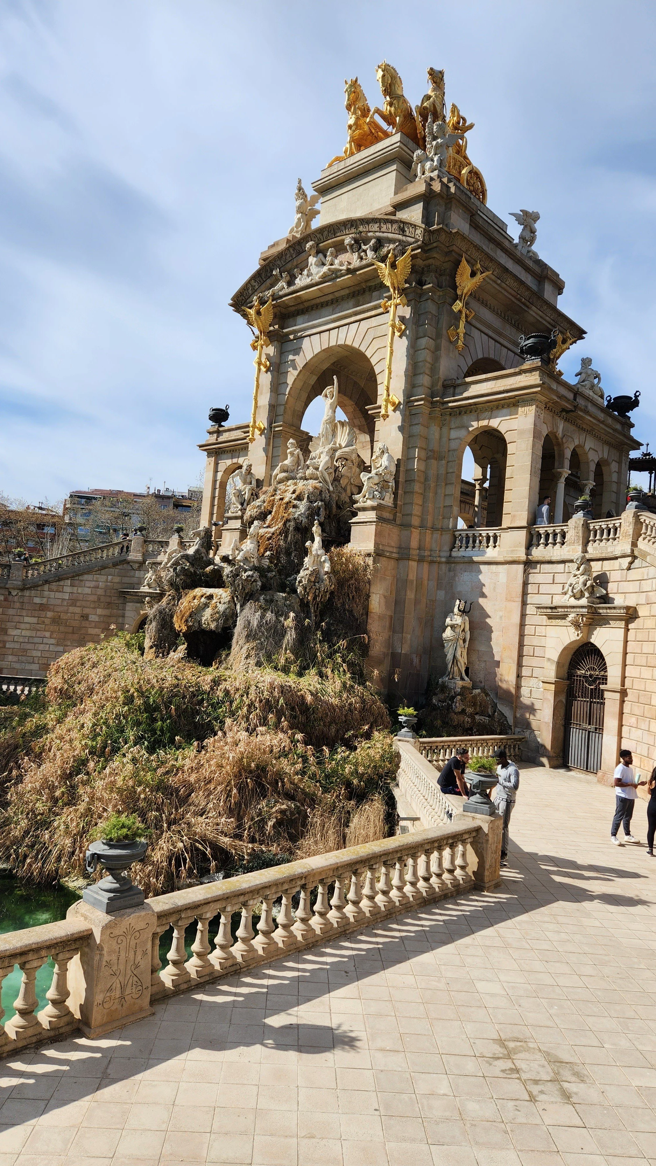 A grand architectural structure with intricate sculptures and golden statues on top. A fountain with water features flows at the base, and there is a terraced area with people standing and sitting, overlooking the structure. The sky is partly cloudy.