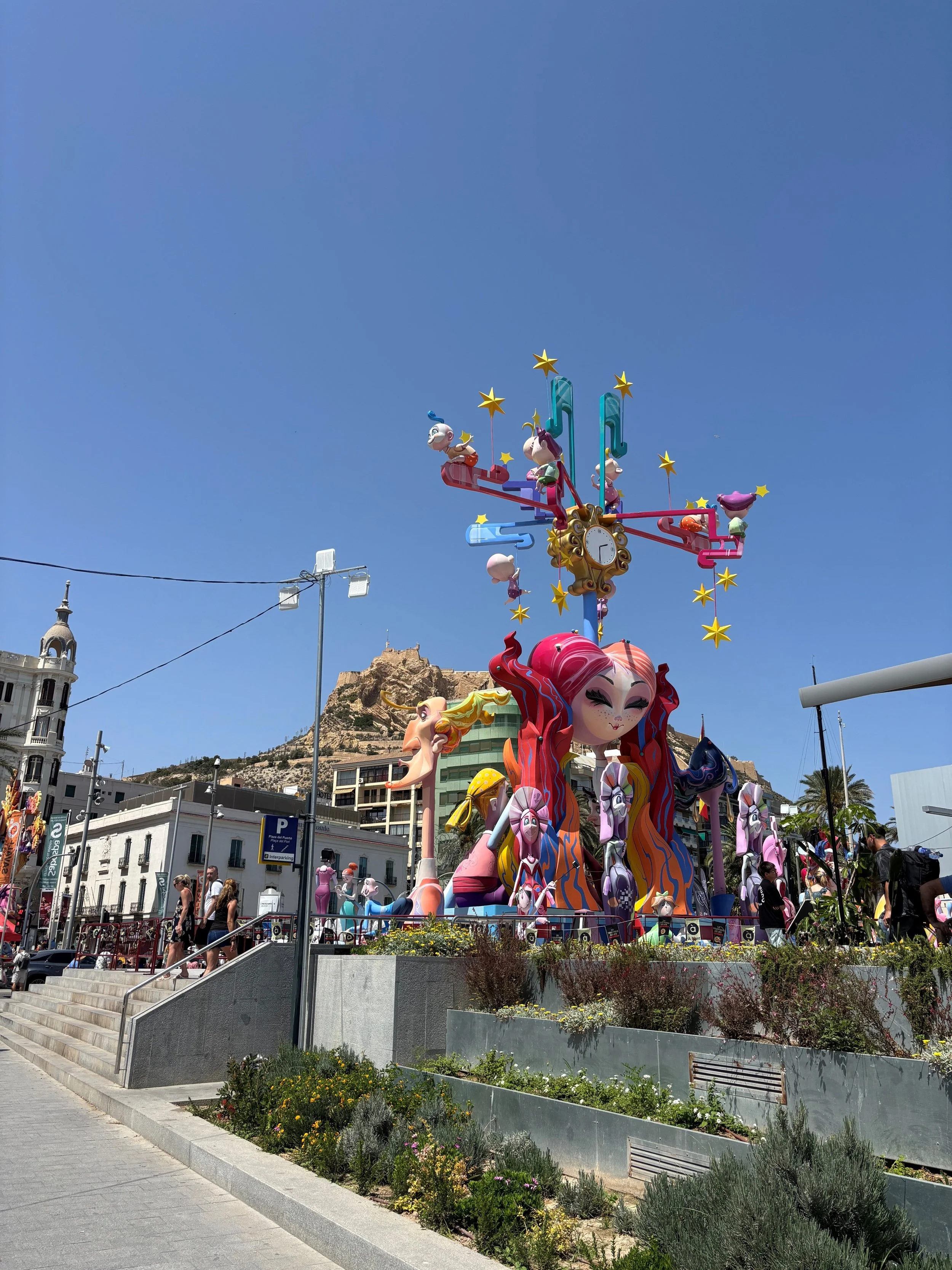 Colorful festival float featuring animated character sculptures, set in an urban area with buildings, stairs, and a mountain in the background on a sunny day.