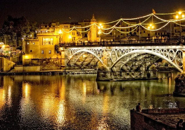 Night view of a bridge over a river with illuminated buildings and string lights in the background, reflecting in the water.