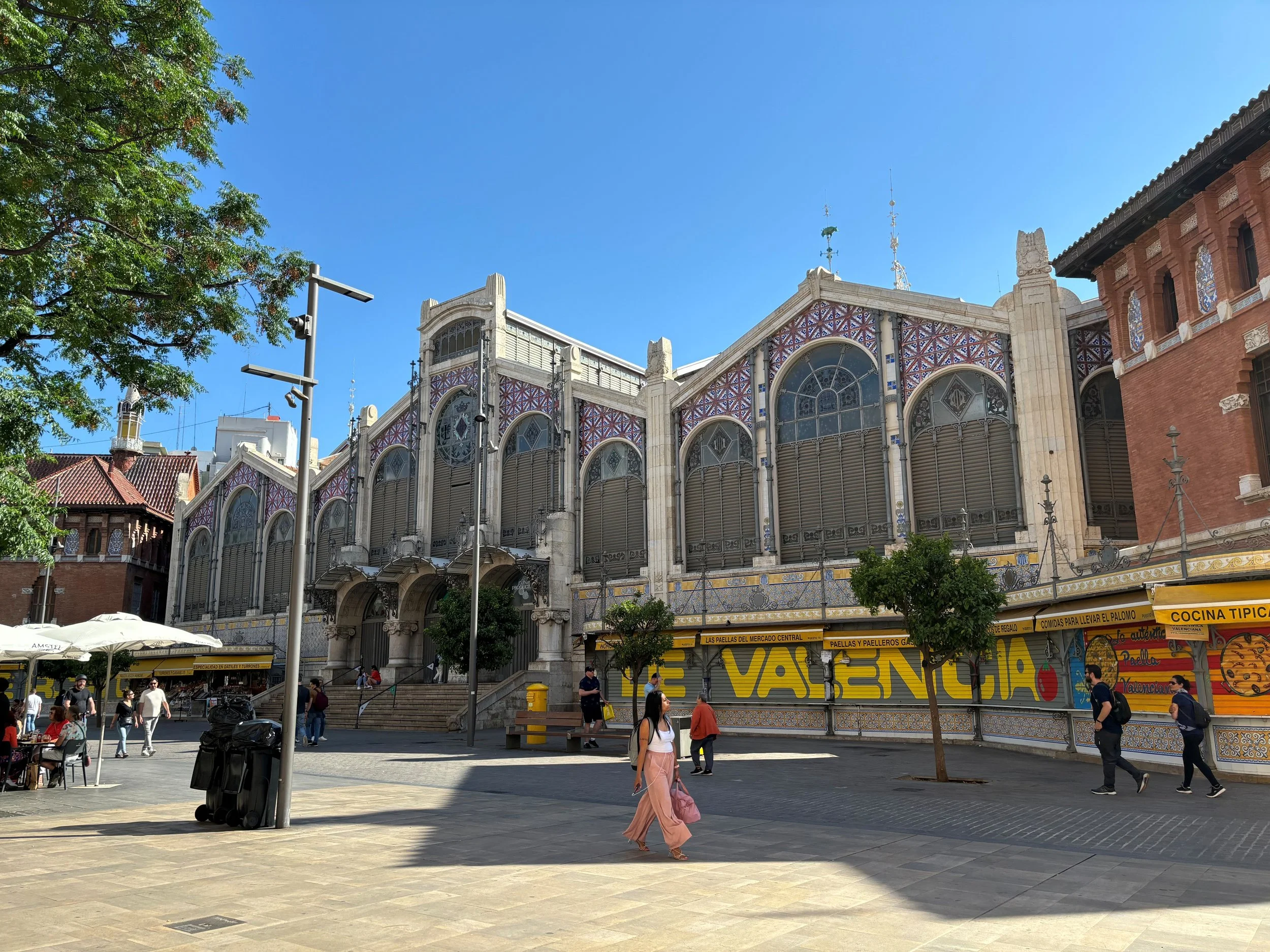 A busy outdoor plaza with people walking and sitting at tables, surrounded by historic buildings with detailed architectural features and a clear blue sky above.