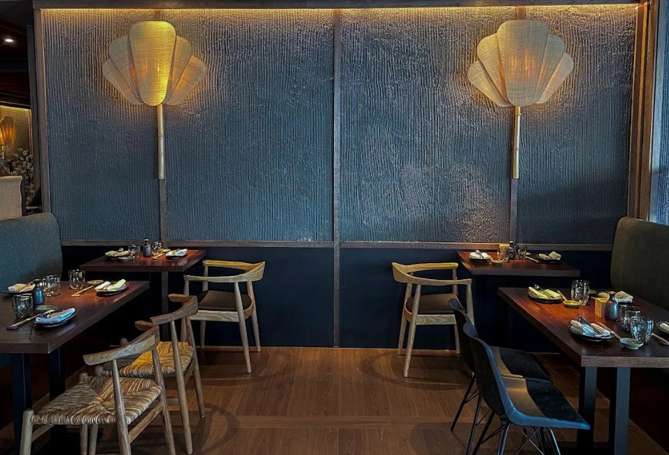 Interior of a restaurant with dark wood tables set with plates, glasses, and utensils, blue upholstered banquettes, and wall sconces shaped like fan lotus flowers.