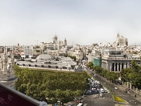 Cityscape view of Madrid, Spain, with historic buildings, green trees, and a busy street filled with cars.