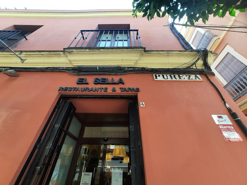 Exterior of a restaurant and tapas bar called El Sella, with a pinkish building facade, balcony, and street signs that say Pureza and the number 4.