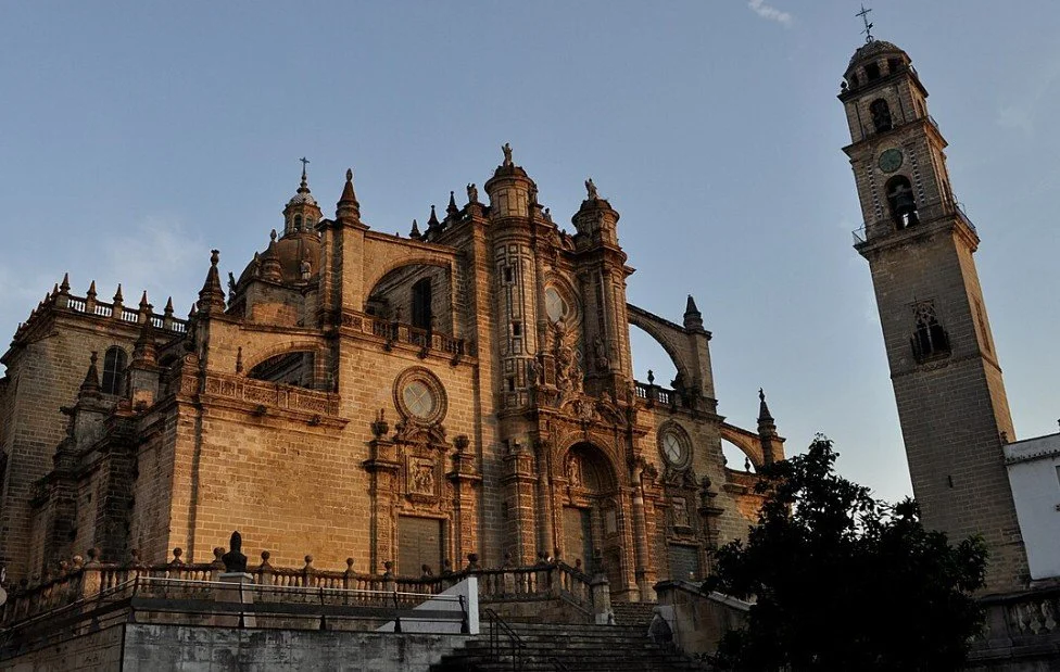 A historic stone cathedral with multiple ornate towers and a nearby clock tower, set against a clear sky.