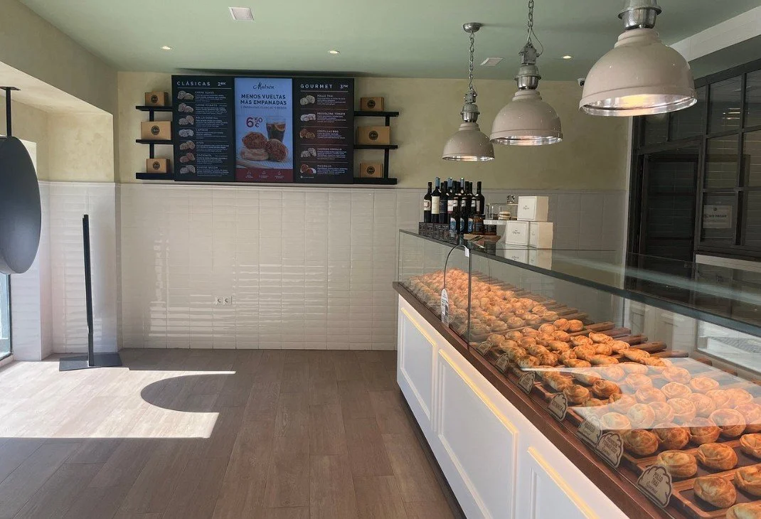 A bakery display case filled with croissants in a modern bakery or cafe with hanging lights, bottles of wine on the counter, and a digital menu board on the wall.
