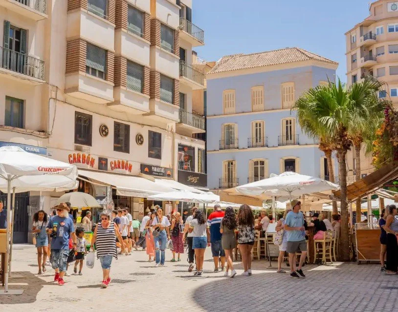 People walking and dining at outdoor cafes on a sunny street with tall buildings, umbrellas, and a palm tree.