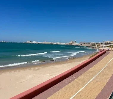 Beach with sandy shore, ocean waves, oceanfront walkway, and cityscape in the distance under a clear blue sky.