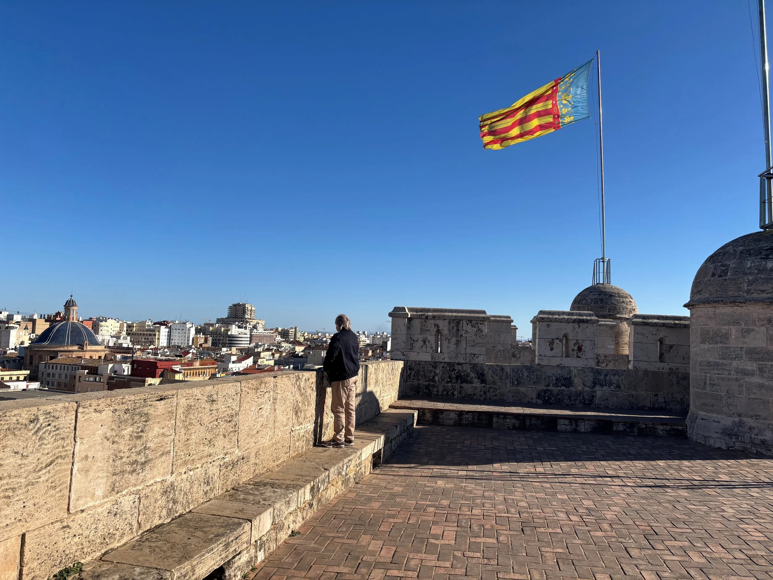 A man standing on a stone and brick balcony overlooking a cityscape under a clear blue sky. A large flag with yellow and red stripes and a blue section with gold details is flying on a flagpole.