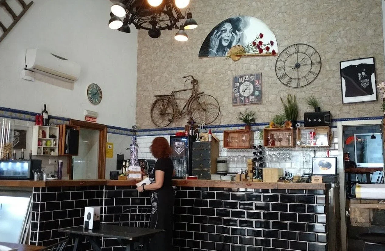 Interior of a cozy cafe with a bar counter, wall decorations including a bicycle, a large clock, wall art, and plants, with a woman standing behind the counter.
