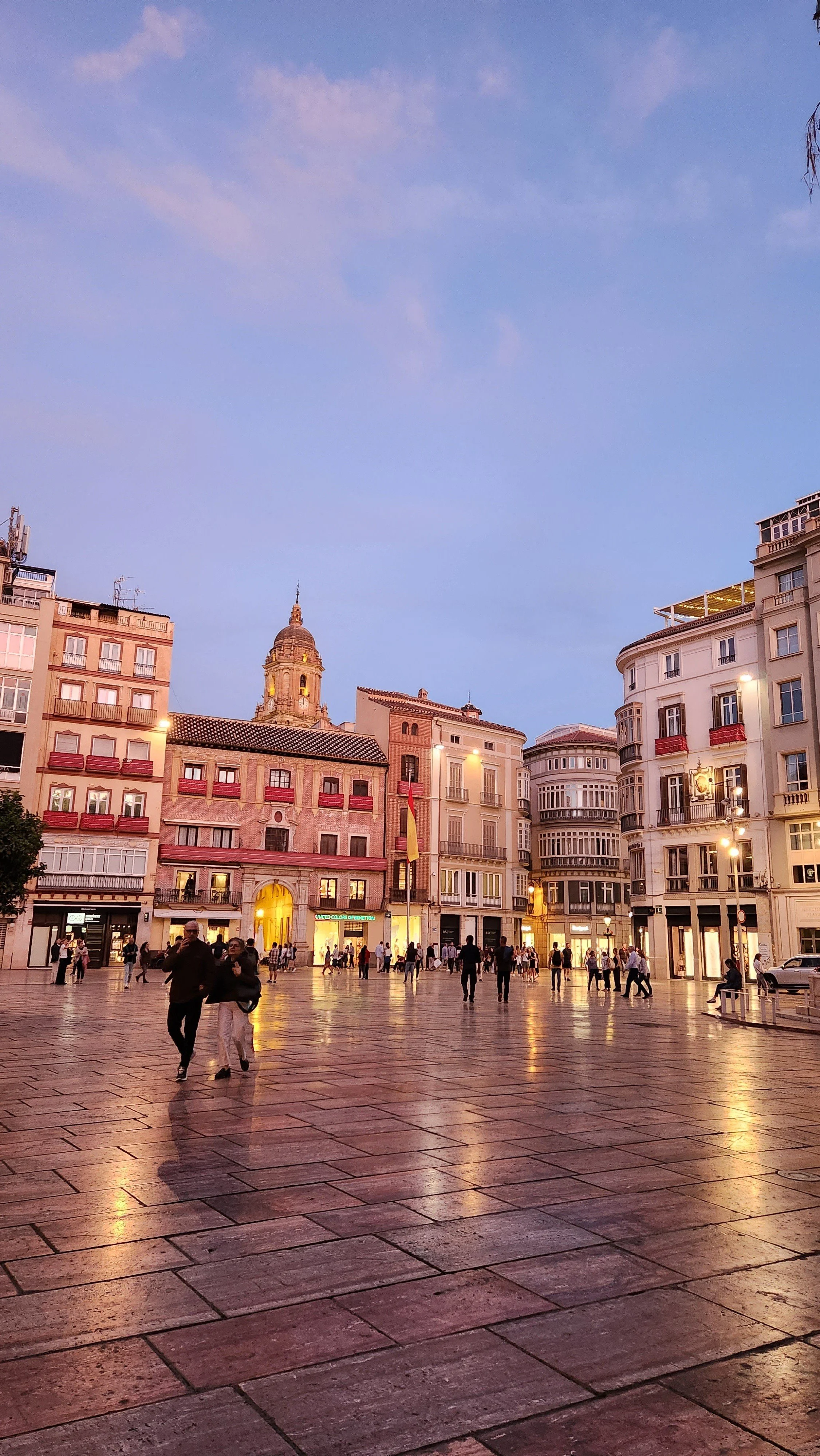 City square at dusk with historic buildings, a prominent dome, and people walking on wet pavement.