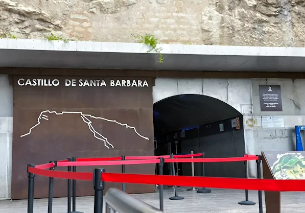 Entrance to Castillo de Santa Barbara with a line diagram of the castle on a brown background, red queue barriers in front, and informational plaques on the wall.