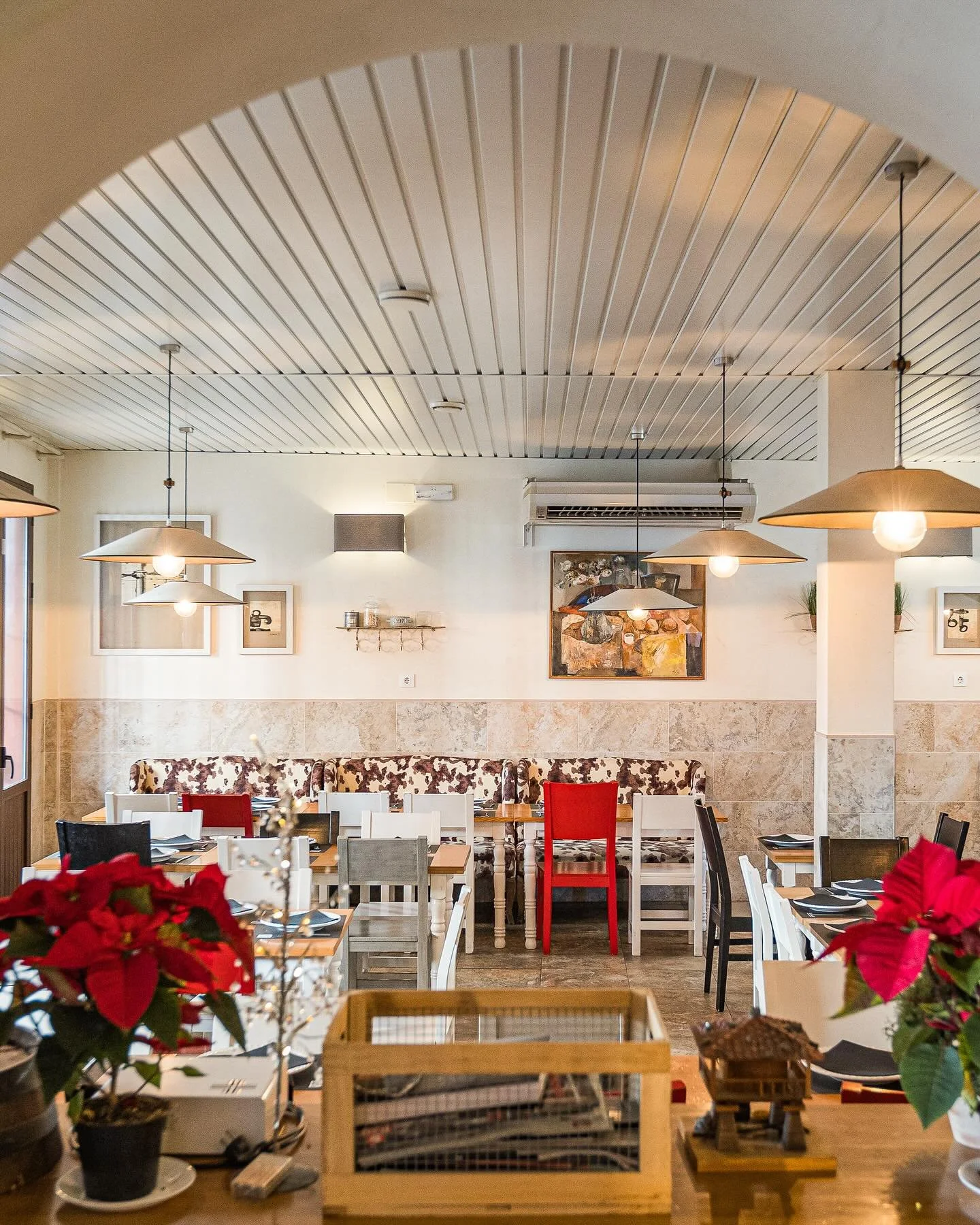 Interior of a cozy restaurant decorated with poinsettias, featuring hanging pendant lights, framed artwork, a patterned sofa, and a variety of chairs and tables.