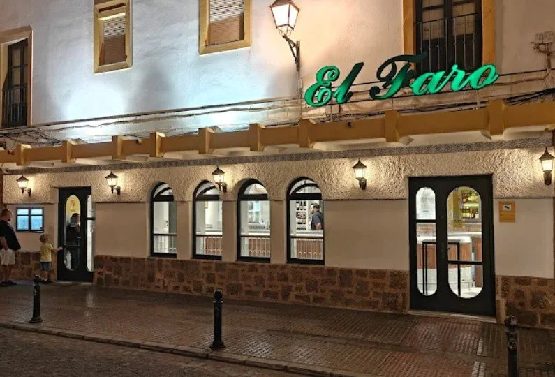 Nighttime view of a building with a sign that reads 'El Faro' illuminated in green. The building has a textured white upper wall with small windows and a lower stone wall. The street in front has bollards and a person with a child standing near the door.