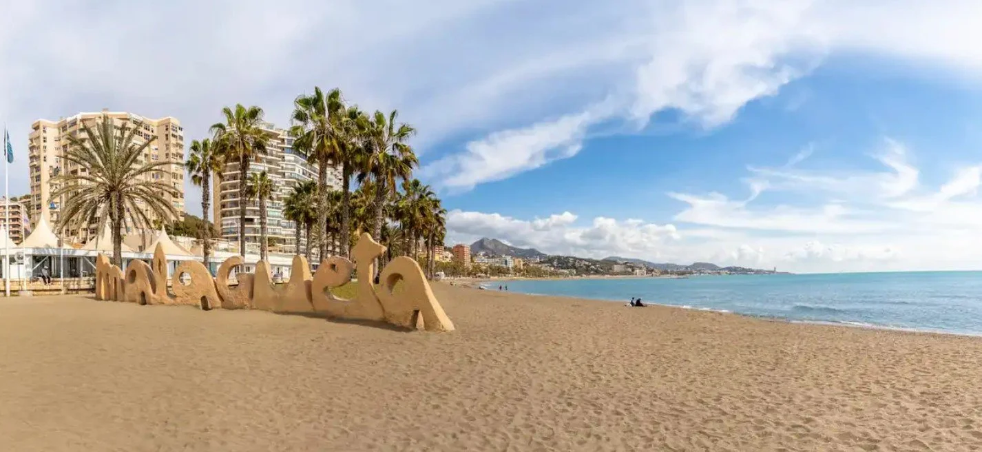 Beach with sand, palm trees, and high-rise buildings in the background. Large sand sculpture spelling 'Malaga' in the foreground.