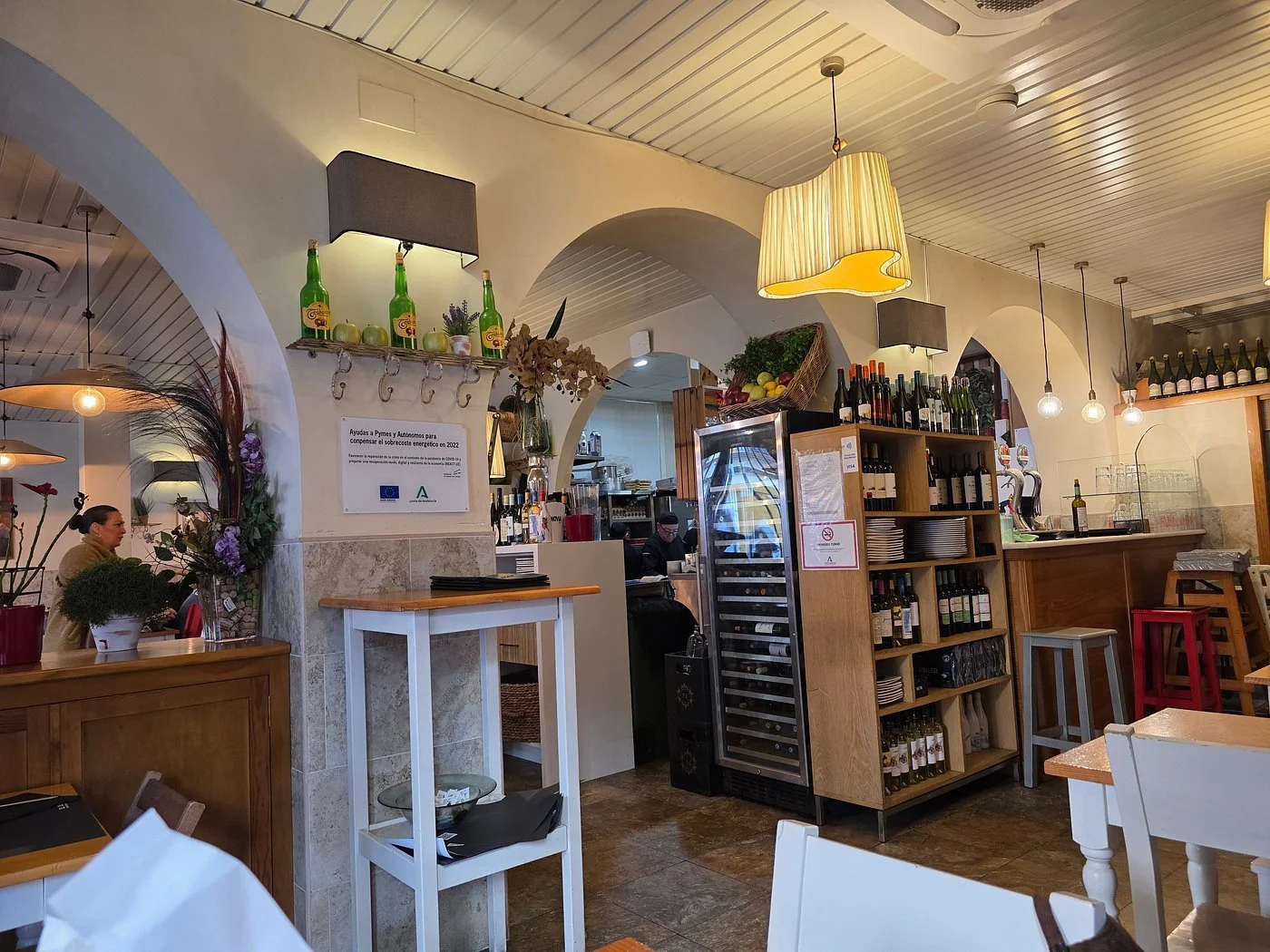 Interior of a cozy restaurant with wooden decor, bar area with wine bottles, tables with white chairs, decorative flowers, and warm lighting.