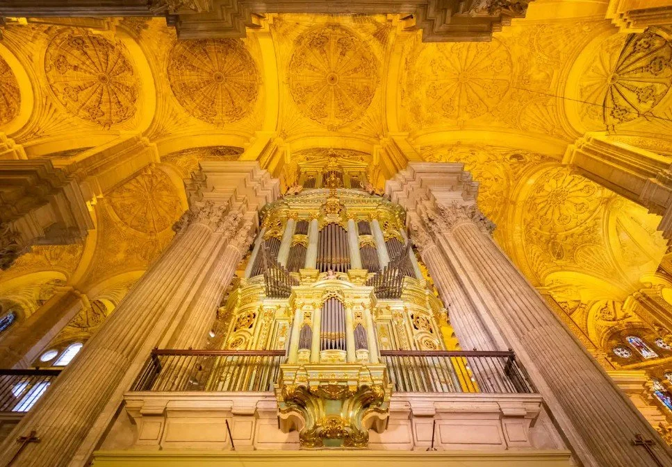 Inside view of a grand ornate church ceiling with a large pipe organ in the center, featuring columns, gold decoration, and intricate artwork on the ceiling.
