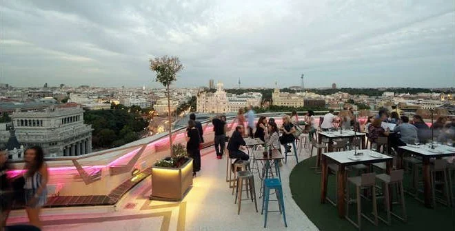 People dining and socializing on a rooftop terrace with a city skyline in the background, featuring a modern lounging area with pink lighting accents, tables, chairs, and a decorative tree.