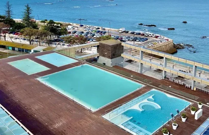 Rooftop view of three pools, including a hot tub, with ocean and parking lot in the background.