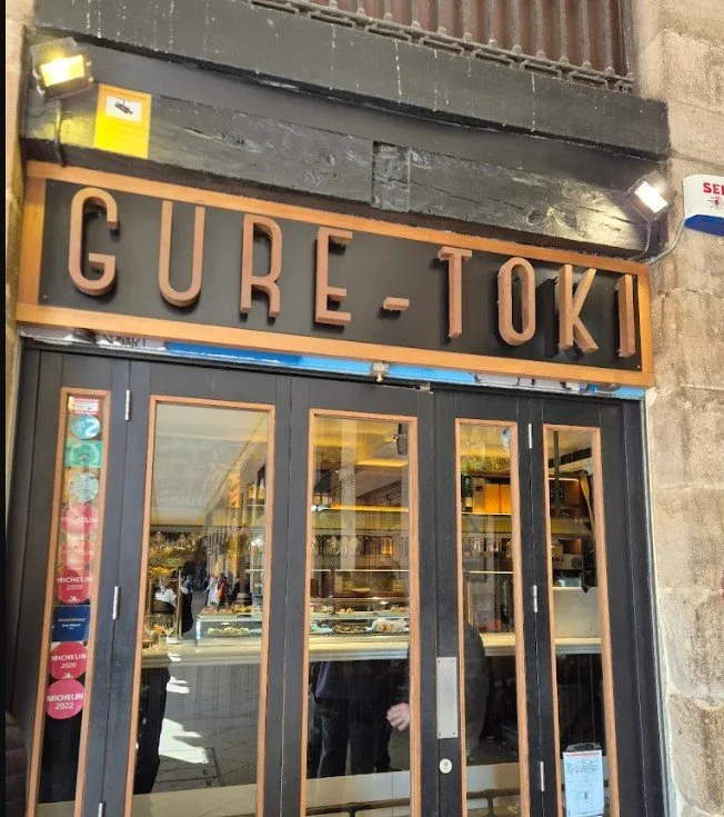 Exterior view of a bakery or café named GURE-TOKI with large glass doors, displaying the interior with shelves and people inside, and visible stickers indicating awards or recognitions.