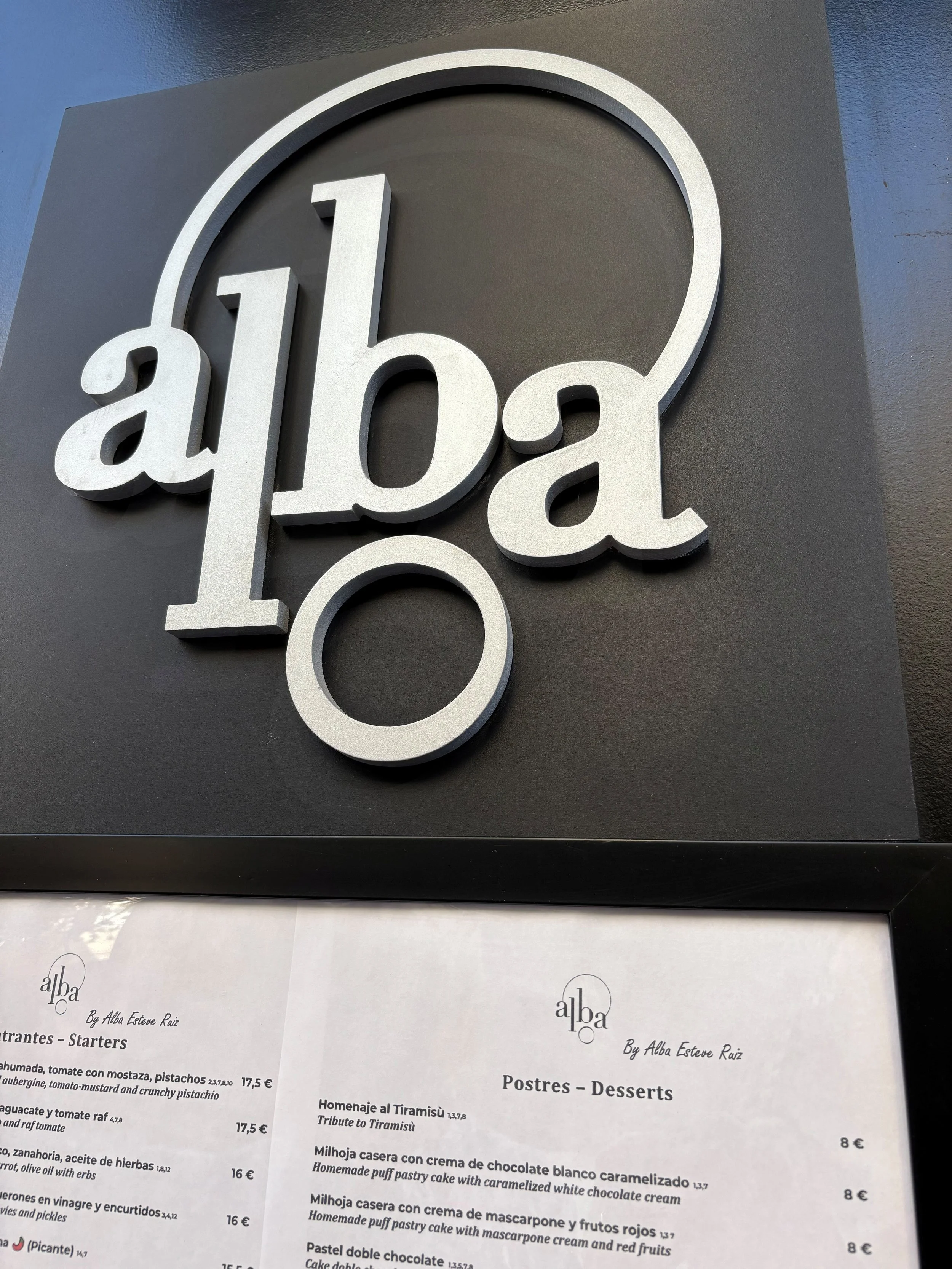 Close-up of a modern black and white restaurant sign with the word "alba" in large white letters, mounted on a dark wall.