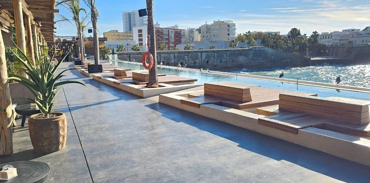 Empty poolside seating area with wooden benches and lounging chairs along a waterfront with city buildings in the background on a sunny day.