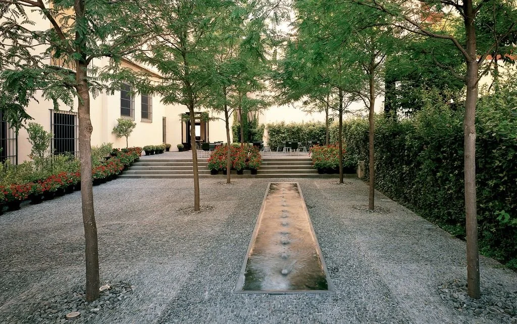 A landscaped outdoor courtyard with a narrow water feature, trees lining both sides, and steps leading up to a seating area with tables and chairs.