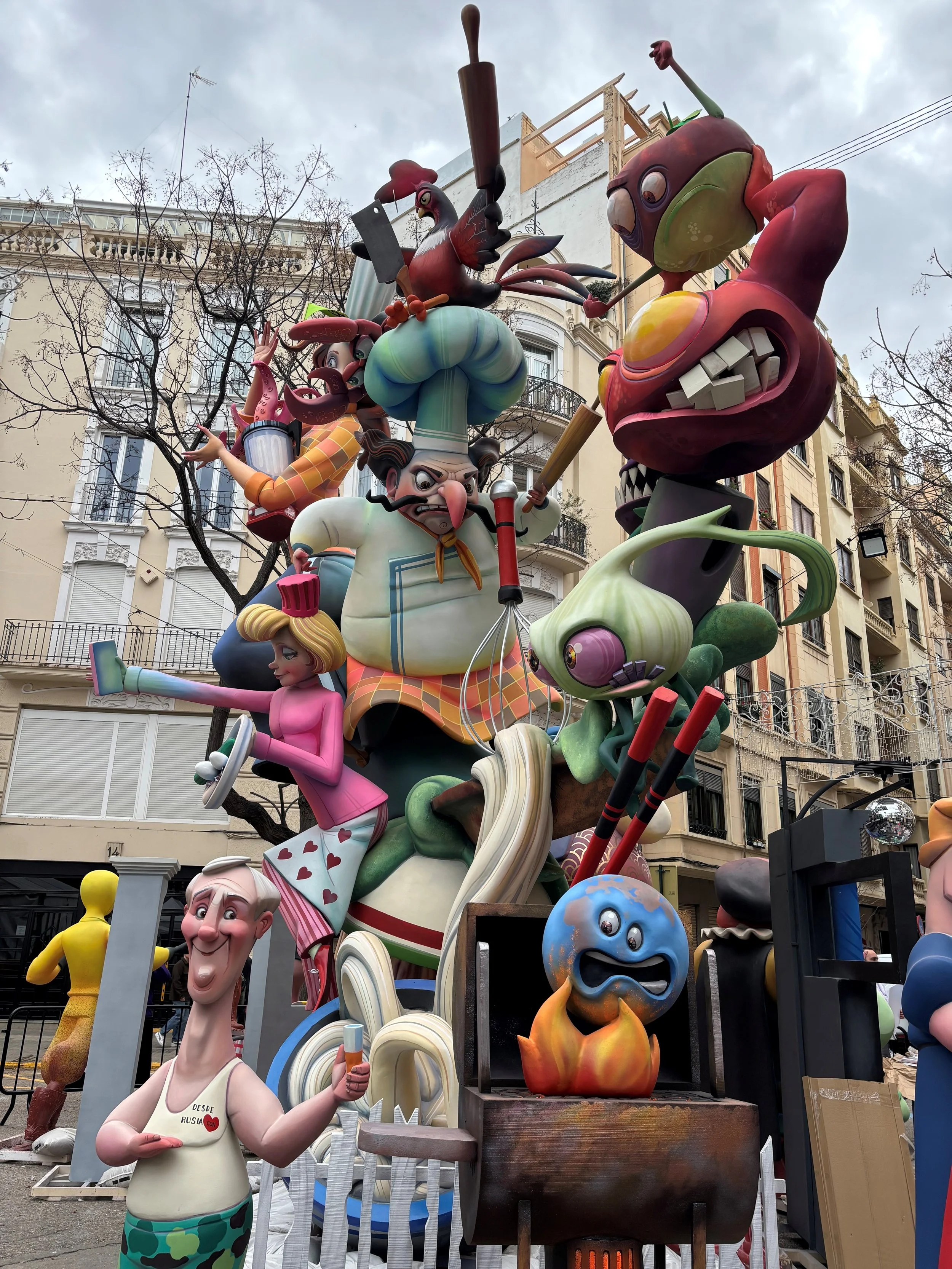 Colorful sculpture of cartoonish characters including a chef, a person with a hammer, a large cactus, a person with glasses, and various imaginative creatures, displayed outdoors against a backdrop of buildings and leafless trees.