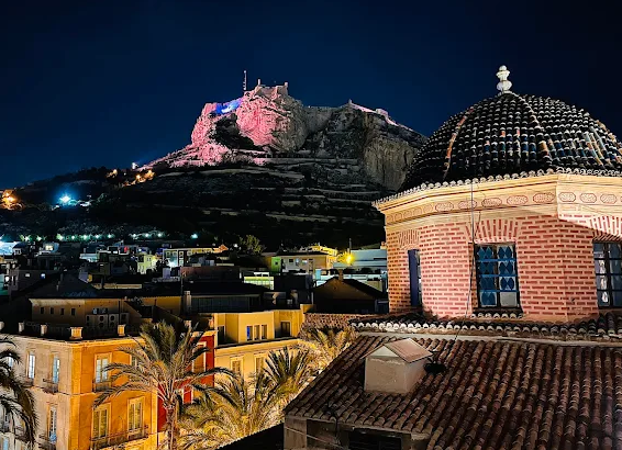 Nighttime cityscape with colorful buildings, palm trees, a brick building with a domed roof in the foreground, and a hilltop fortress illuminated in the background.