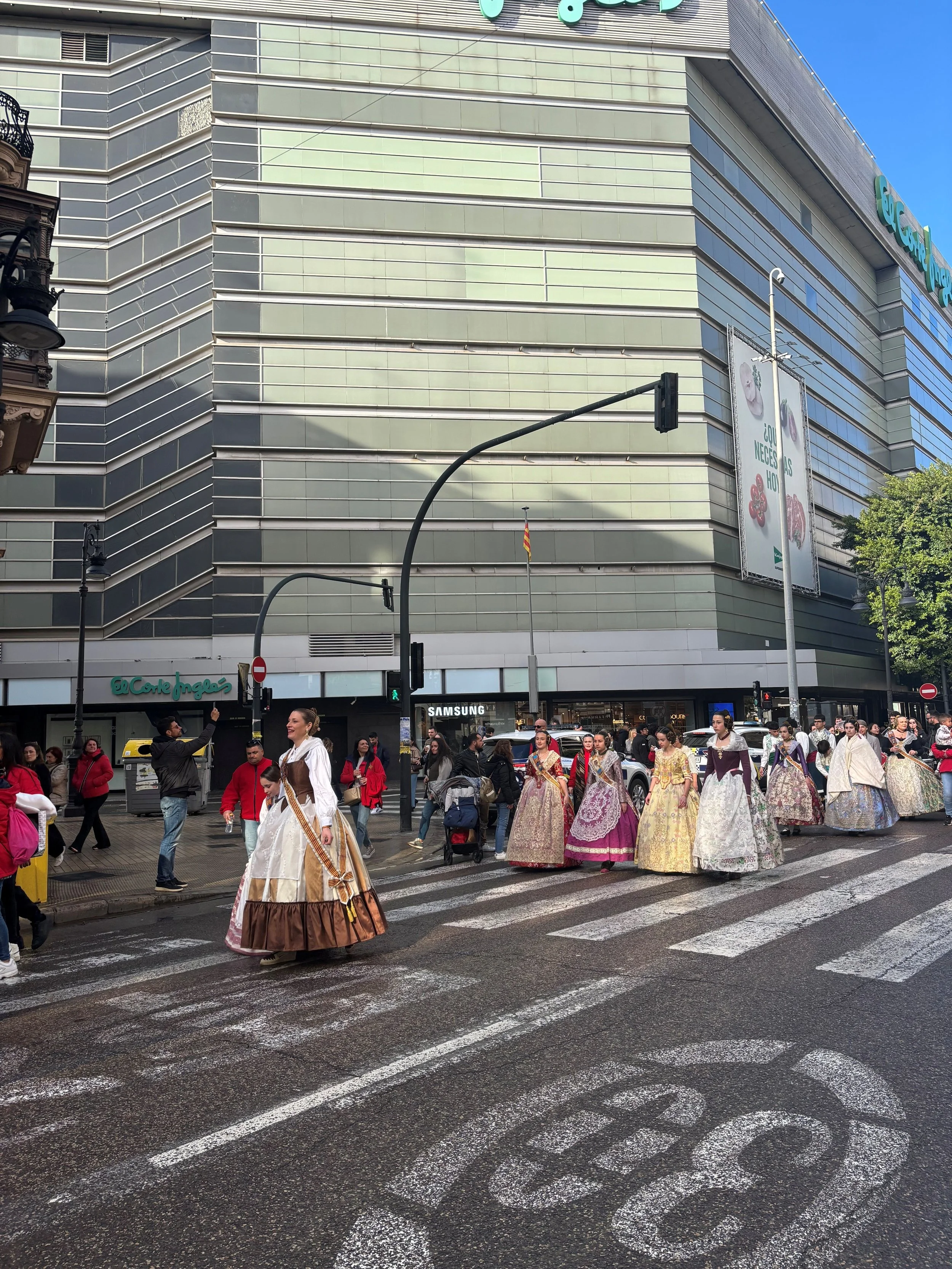 People dressed in traditional European costumes walking across a city street with a modern building and street signs in the background.