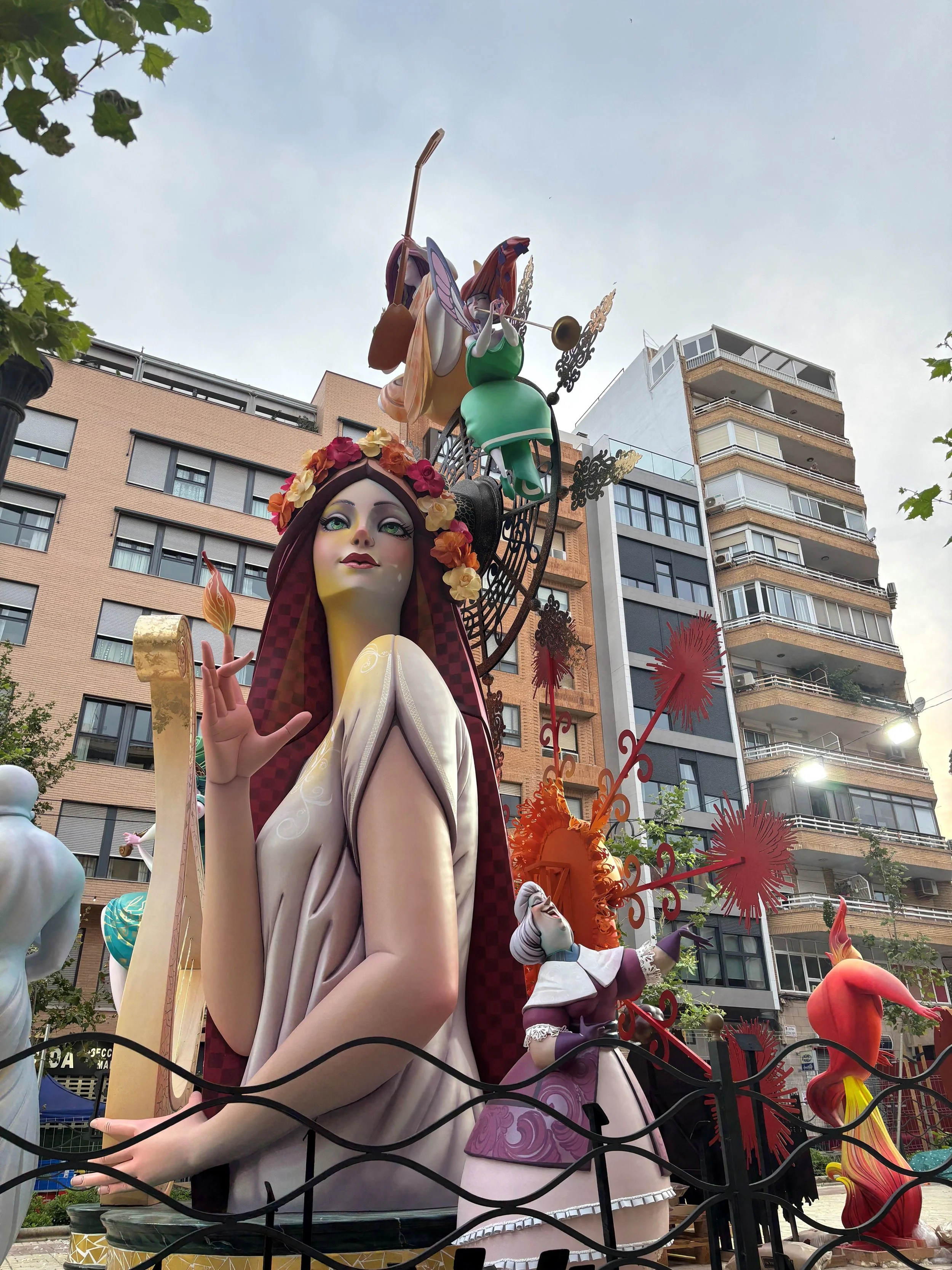 Large colorful sculpture of a fairy with long brown hair, green dress, and purple wings, playing a trumpet at a street parade with buildings in the background.