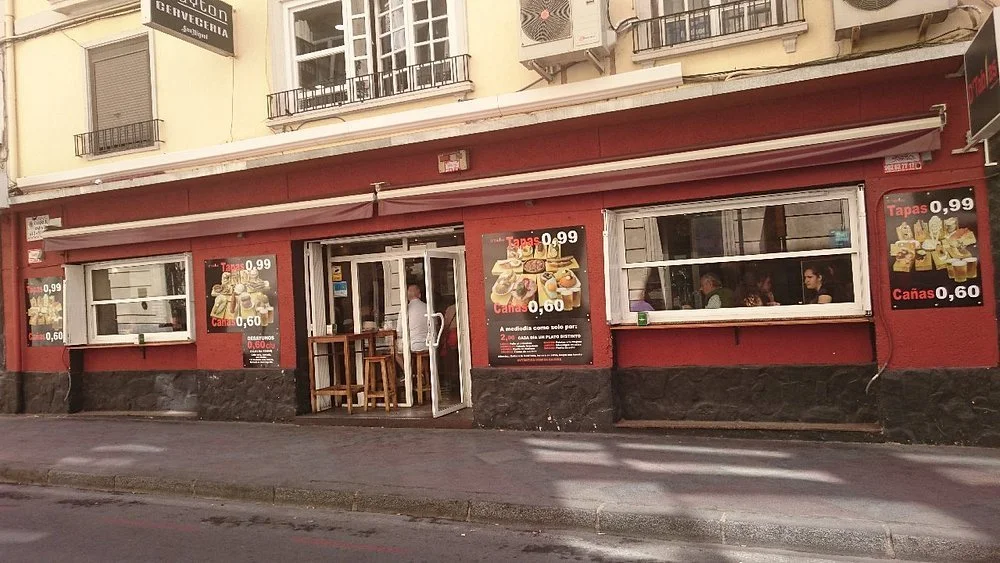 Exterior of a small restaurant or cafe with a red facade, open entrance door, and three large windows showing patrons inside. Several posters advertising tapas and drinks are on the outside walls, with prices listed. There are outdoor wooden tables and stools near the entrance.