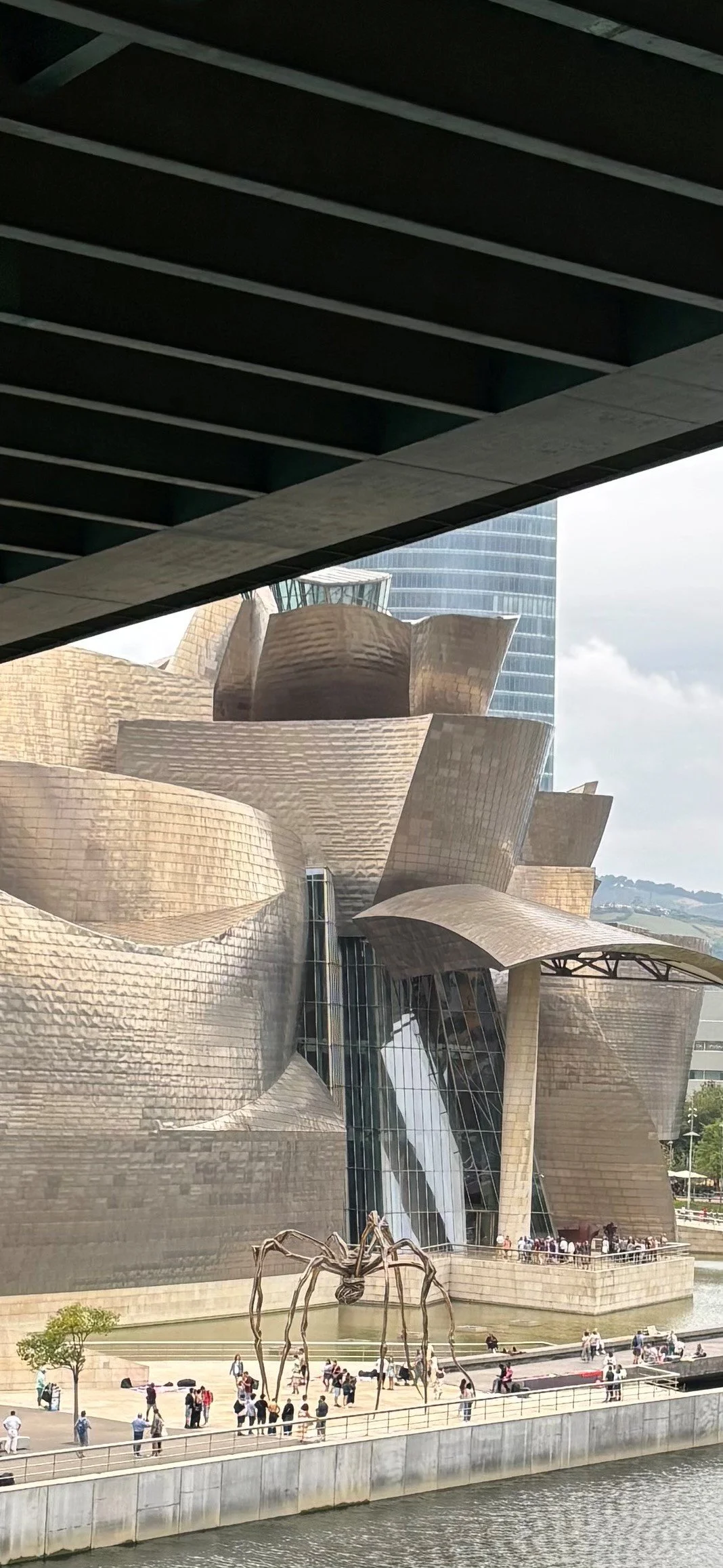 The image shows the Guggenheim Museum Bilbao, a modern architectural building with curvy metallic surfaces, situated beside a water body with a large spider sculpture and people gathered in the area.
