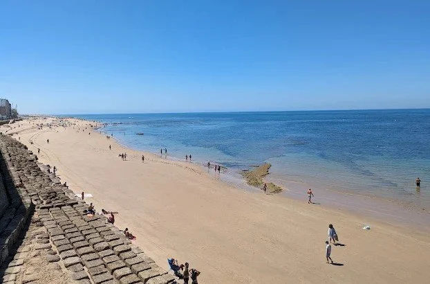 View of a sandy beach with people walking and relaxing, clear blue sky, calm ocean waves, and a stone wall along the shoreline.