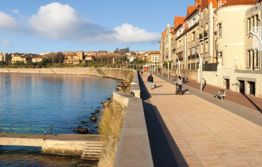 People walking along a waterfront promenade with benches and streetlights, surrounded by historic buildings and a calm body of water with rocks and a small bridge.