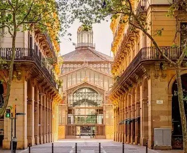 A covered walkway between two yellow buildings with balconies, leading to a glass-domed structure at the end, surrounded by trees.