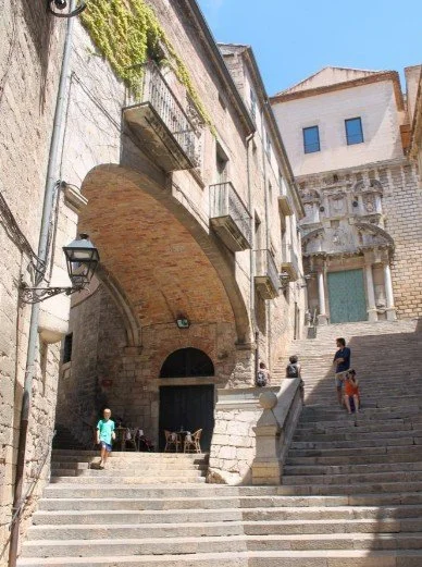 Stone stairs leading up past historic buildings with balconies and an archway, with a few children walking and sitting outside.