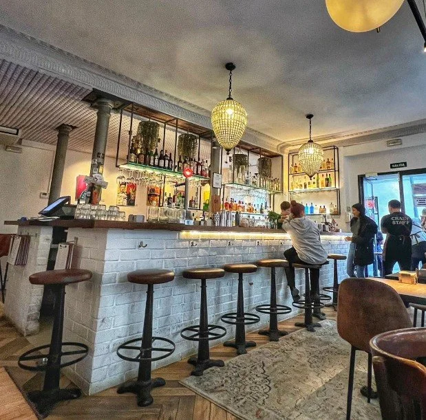Interior of a bar with a white brick counter, barstools, hanging pendant lights, and shelves with bottles and decorative plants behind the bar. There are patrons sitting and standing near the counter, and a door with an exit sign on it.