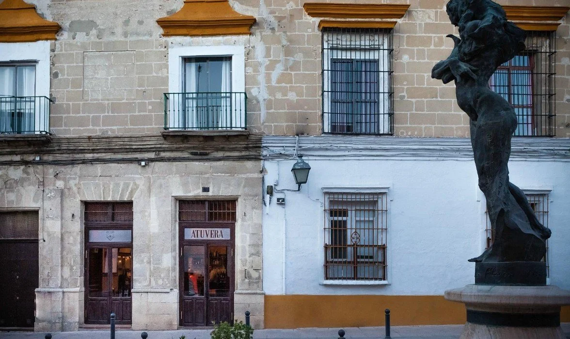 A street scene featuring a stone building with three large windows, each with wrought iron bars, and a door with the word "ATUVERA" on it. There is a statue of a woman with flowing hair on a pedestal to the right.