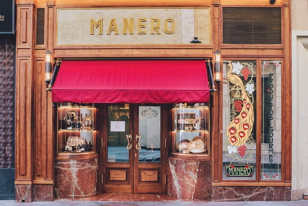 Facade of a bakery or pastry shop named Manero with wooden framing, red awning, rear-lit display windows showing baked goods and bottles, a decorative window with a peacock design and grapevine, and the words 'Productions Gourmet' on the glass.