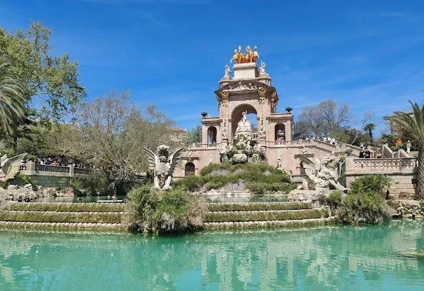 Historical monument with ornate architecture and sculptures surrounded by gardens and a water feature, set against a clear blue sky.