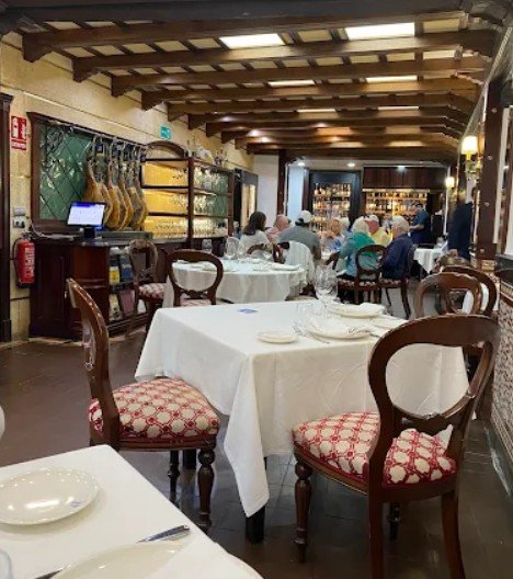Interior of a restaurant with white-clothed tables, wooden chairs with patterned red and cream upholstery, and a display of hanging cured meats.